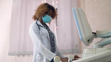 Close-up of face of professional african american obstetrician gynecologist in protective mask performing ultrasound of belly of pregnant woman. Concept of medicine, health care and people video