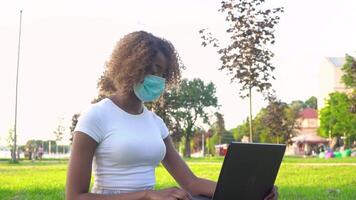 Young african american female student in protective mask working on laptop in park during covid 19 pandemic video