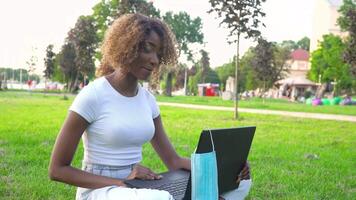 giovane africano americano donna chiamata su il computer portatile parla con in linea insegnante studiando nel il città parco durante covid 19 pandemia. medico monouso maschera sospeso su Schermo video