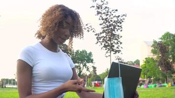 Young african american woman calling on laptop talks with online teacher studying in the city park during covid 19 pandemic. Medical disposable mask hanging on display video