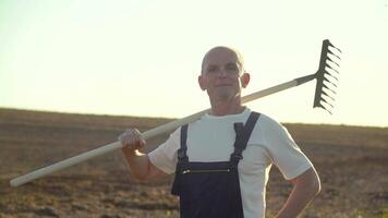 A senior farmer standing through a field with rake. Farming and agriculture concept video