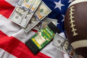 Online sports betting. Dollars are falling on the background of a hand with a smartphone and a soccer ball. Creative background, gambling photo