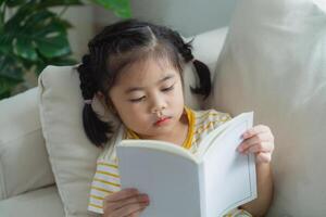 alegre contento asiático niño bebé niña concentrado y leyendo libro mientras sentado en sofá sofá en vivo habitación a hogar. niña relex leyendo libro sonrisa a sofá en el casa. espalda a colegio concepto. foto