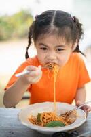 joven niña niños disfrutando un tenedor de espaguetis. de cerca de un pequeño niña en un naranja camisa comiendo espaguetis con un enfocado expresión, al aire libre restaurante. foto
