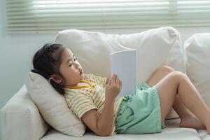 alegre contento asiático niño bebé niña concentrado y leyendo libro mientras sentado en sofá sofá en vivo habitación a hogar. niña relex leyendo libro sonrisa a sofá en el casa. espalda a colegio concepto. foto