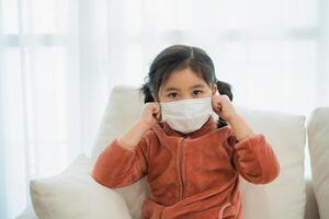 Young Asian Girl Adjusting Her Protective Face Mask. A young child in a cozy fleece adjusts her face mask while sitting on a sofa, looking directly at the camera. photo