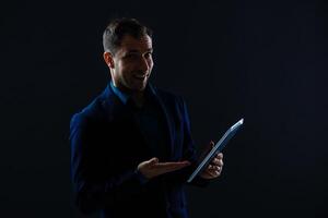 un grave hermoso joven empresario en un traje utilizando un tableta en frente de un oscuro gris antecedentes en un estudio. foto