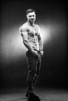 Full size photo of an athletic guy posing with hands in pokets. Studio shot. Bodybuilder. Dark grey background. Closeup.