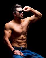 Well build handsome man without a shirt posing in studio. Strong body and cool face. Male wearing glasses, sitting on a chair with hands on legs. selective focus. Closeup. photo