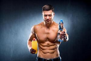 Shirtless athletic muscular guy holding bottles with still drinks in hands on the dark grey bachground. Half waist photo. Portrait. Closeup photo