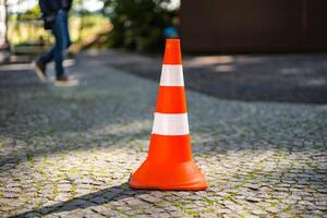 Plastic orange parking cone standing in the street on the blurred background of male's legs. Striped road sign for parking in the pavement. Close-up photo