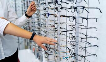 Row of glasses at an opticians. Eyeglasses shop. Stand with glasses in the store of optics. Woman's hand chooses spectacles. Eyesight correction. photo