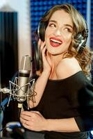 Attractive girl singer with headphones in front of the microphone sings with mouth wide open and with an expression of happiness on her face. Young woman singing in recording studio. photo