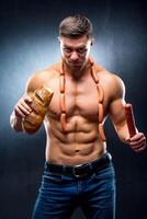 man athlete with naked torso in a black background holds bread in one hand and smoked sausage in the other hand with sausages on a neck. lifestyle concept. photo