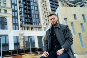 Young businessman sitting on branch at the street. Tourist have a rest after long travel. Handsome stylish man waiting his transport. photo