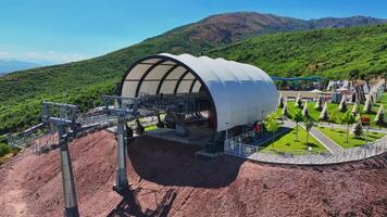 The drone flies over the mountain cableway station. It's a sunny summer day, surrounded by hills covered with greenery. video