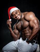 africano americano hombre en Navidad sombrero. atractivo hombre con desnudo abdominales sesión. Papa Noel claus sombrero. foto