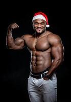 hermoso muscular hombre en Navidad sombrero demostración su bíceps. atlético aptitud Papa Noel claus con desnudo torso. foto