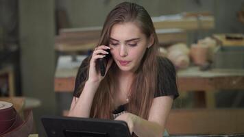 Cheerful young carpenter discussing order details with her customer on smartphone and taking necessary notes, interior of spacious workshop on background video