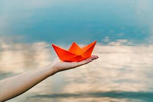 Nice red paper boat on the hand of a young woman above the water in the river background. Woman holding origami boat near river. Close-up photo