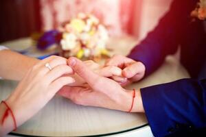 Bride and groom holding hands with wedding rings on the table over the blur bouquet background. Wedding couple holding hands. Close-up photo