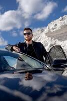 Attractive man standing near opened car door. Nice view of mountain and sky. Stylish combination of black and white. photo