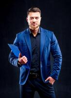 Half-length portrait of handsome young smiling man wearing dark shirt and blue suit holding blue folder, looking at camera. Black background. Money and business concept. photo