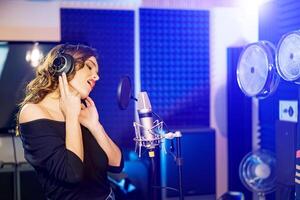Side view of a pretty woman standing in studio with headphones and singing in front of the microphone. Recording studio in blue colors and the young lady singing. photo