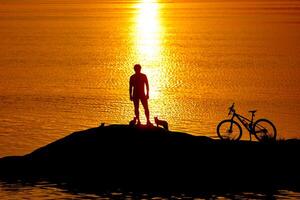 silhouette of a cyclist at sunset with a blurred reflection in the water with ripples. Rest on rocks. Standing near water. photo