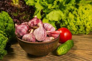 A full bowl with garlic bulbs. Tomatoes and cucumbers on a wooden table. Green salad in the background. photo