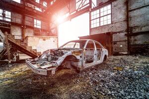 Metal frame of a rusty car in a big old abandoned building inside with light leaks. Broken and forgotten rusted car frame on the background of ruined plant photo