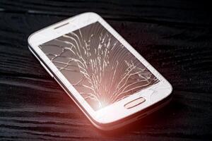 Damaged screen of a broken smartphone isolated on dark background. Crashed mobile phone lying on the floor inside. Close-up photo