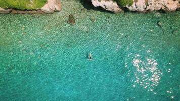 einsam Frau genießt makellos Türkis Gewässer. Antenne oben Nieder Aussicht von Mädchen Schwimmen im still Ozean mit felsig Küste im das Distanz. video