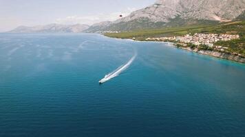 vedette sculpte une mousseux Piste dans le calme adriatique mer. au-dessus de, une rouge parachute ascensionnel flotteurs en dessous de une clair ciel. video