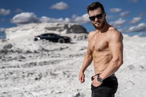 Fashionable guy with naked abs standing in front of dark contemporary car. Bodybuilder in sunglasses and wirstwatch. White mountains and beautiful sky. photo