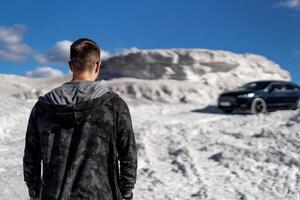 Serious man standing with his back to screen. Dark car on background. White mountain and beautiful view. photo