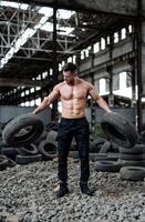 Sportive man holding heavy tires. Handsome man with naked abs in jeans. Abandoned house on background. photo