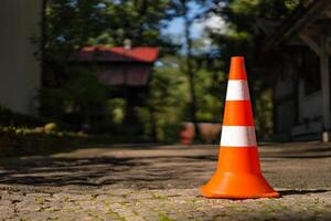 Red orange cone with a white stripe on the paving stone road. Drive safety and constructions concept. Closeup. Blurred background with green trees and buildings. Sunlight and shadow. photo