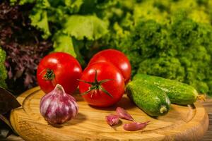 A wooden board with fresh tomatoes, garlic and cucumbers. Green salad in the background. photo