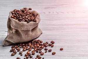 Small sack of coffee beans and some grains lying near it on the light background. Coffee beans scattered near the bag over the wooden table. photo