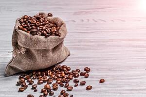 Small sack of coffee beans and some grains lying near it on the light background. Coffee beans scattered near the bag over the wooden table. photo