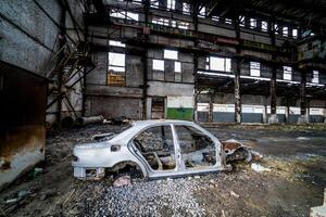 Side view of an old rusty antique car frame on the background of broken-down plant. Interior of the large abandoned building with a damaged car photo