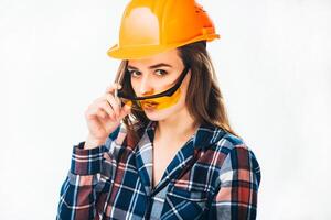 Beautiful builder woman wearing orange protective helmet looks over yellow safety glasses at camera isolated on white background. Close-up photo