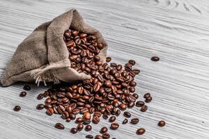 Bag of natural brown coffee beans and some scattered seeds on the light wooden background. Roasted coffee grains spilling out of sack on the table. photo