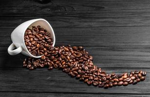 Roasted coffee beans spilling out of a white cup on the wooden table. Natural coffee seeds scattered nicely on grey surface. photo