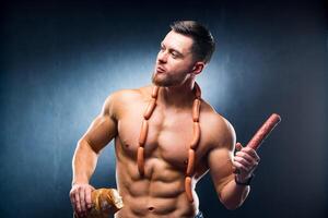 Bodybuilder athlete with naked torso in a black background holds bread in one hand and smoked sausage in the other hand with sausages on a neck. lifestyle concept. photo