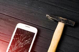 Composition of a hammer and a smart phone with a broken screen over the dark wooden surface. The concept of a device failure photo