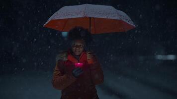 Jeune africain femme permanent sur rue à nuit pendant neige tomber avec parapluie video