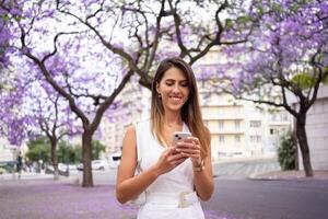 Smiling woman using smartphone near violet Jacaranda trees in city photo