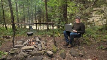 un' uomo lavori su un' il computer portatile vicino un' fuoco di bivacco nel un' bellissimo foresta. concetto di libero professionista, digitale nomade o a distanza ufficio. 4k video